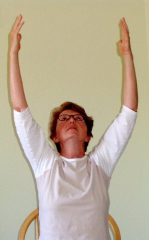 Photograph of an older woman with short brown hair and black framed glasses. She is sitting in a wooden chair with her arms stretched above her head, a shoulder width apart, as she looks up towards the ceiling. She is wearing a white, ¾ sleeve shirt and the wall behind her is beige. 
