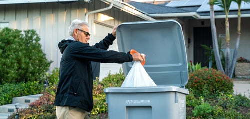 City resident placing a trash bag inside a gray collection bin by the curb