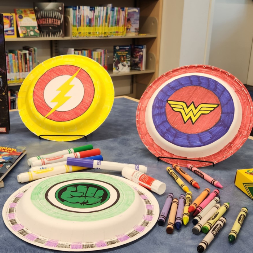 Colorful paper plate super hero shields on a table with markers and crayons.