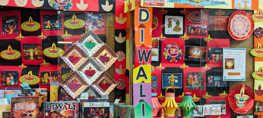 Mira Mesa Library fills two display cases with a black and red checker background that proudly feature Diwali books and diyas in the front. The display cases are lined with black and red squares and orange and yellow squares. The words “DIWALI” are spelled vertically in a rainbow pennant in the middle of the two display cases.