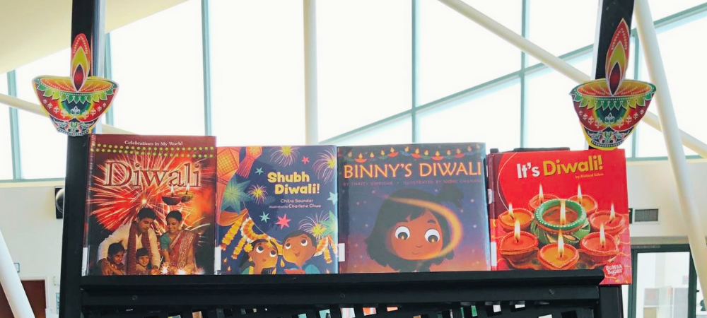 The Mira Mesa Library proudly showcases a Diwali display on a black shelf lined with colorful images of diyas or oil lamps. The shelf portrays four children’s books. The books are titled: Diwali, Shubh Diwali!, Binny’s Diwali, and It’s Diwali!