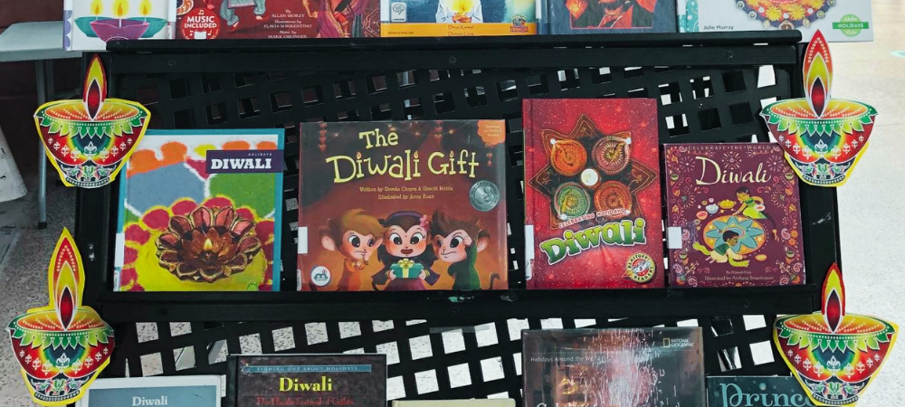 The Mira Mesa Library proudly showcases a Diwali display on a black shelf lined with colorful images of diyas or oil lamps. The shelf portrays four children’s books. Three books are juvenile nonfiction books about the Diwali holiday and one book is a picture book called The Diwali Gift featuring three little monkeys on the cover.