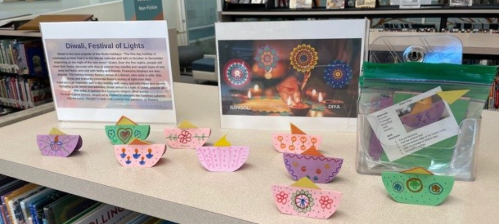 At the Rancho Penasquitos Library, purple, pink, and green paper diyas (oil lamps) line a bookshelf. Behind the paper diyas, there is a sign that provides an explanation about “Diwali, Festival of Lights,” a photograph of rangoli, and container that holds several take-home kits so families can make their own paper diyas at home.