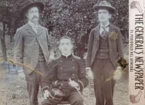 Historical photograph of three men, two standing, one sitting wearing an army uniform with the words “The General’s Newspaper, El periódico del general” along the side