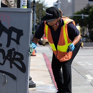 A city worker removing graffiti from an electrical box on a sidewalk