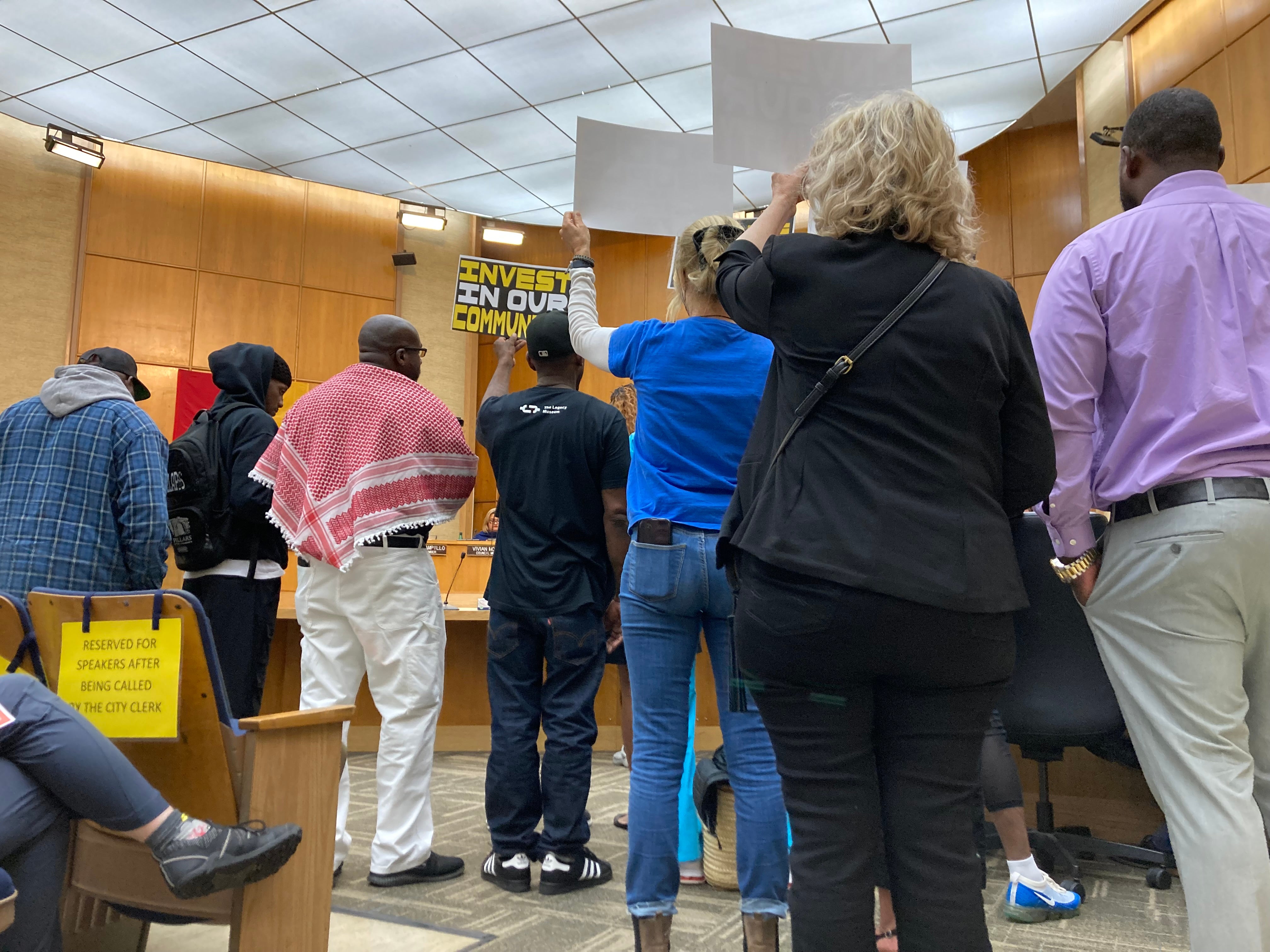 A group stands with signs that say "Invest in our community" at City Council