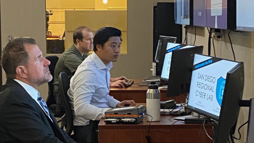 Three people are seated at desks attentively monitoring computer screens displaying the San Diego Regional Cyber Lab emblem, in a professional office environment.