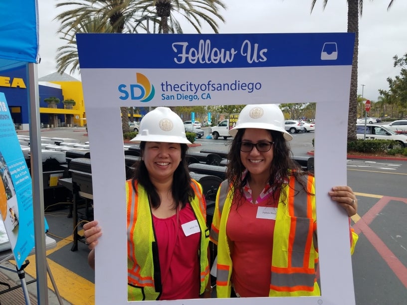 Two smiling women posing inside a handheld frame that says follow the City of San Diego