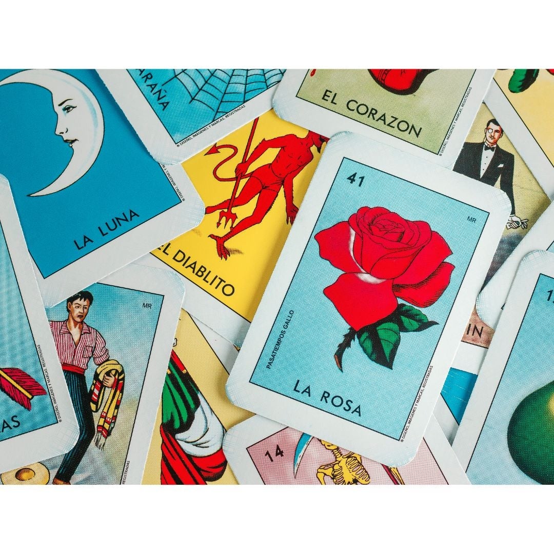 A photograph of a pile of loteria cards. Visible at the top are ”La Rosa” a blue card with a red rose, ”La Luna” a darker blue card with a crescent moon, and ”El Diablito” a yellow card with a red devil. 