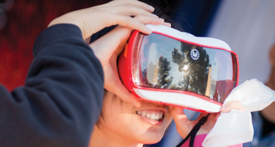  A youth smiling while looking through a red and white viewer with a reflective lens. An out of frame student helps hold the viewer steady. They appear to be outside. 