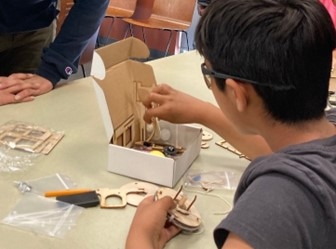 An over-the-shoulder angle of a youth with short black hair and black glasses, wearing a dark gray t-shirt and working on a wood-cut robotics project. He holds assembled pieces of the project in his left hand and is reaching for more parts in a white box with his right hand. There are more pieces and clear plastic bags of smaller components on the table around the box. Another person watches just out of frame.
