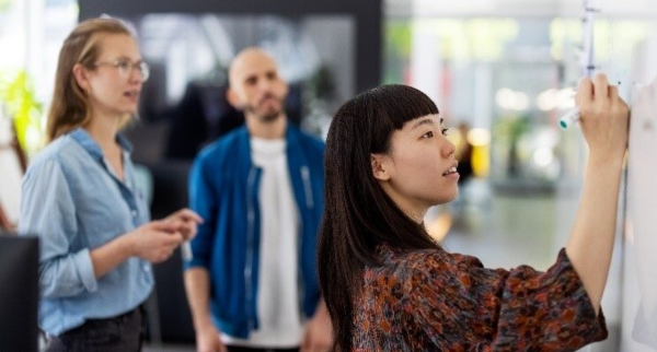 Asian appearing female adult with long black hair collaborating with a male appearing and a female appearing adult looking on in the background, while writing on a whiteboard.