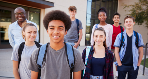 A group of diverse teenage male and female students wearing backpacks and their teacher smiling for the camera. They are outside a building.