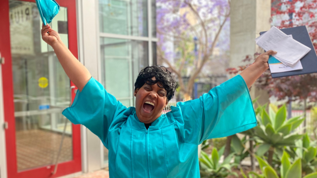 African-American woman with short curls wearing a teal colored graduation gown shouting with excitement with arms raised over her head holding a teal graduation cap in her right hand and graduation program, diploma cover and her commencement speech in her left hand.  She is standing in the library courtyard while behind her there are green plants on right and the glass door outlined in red metal that leads into the library auditorium on the left.