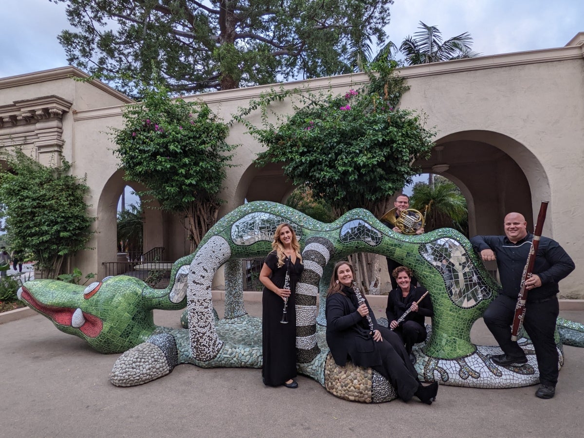 Five people holding instruments standing around a green animal structure in Balboa Park.