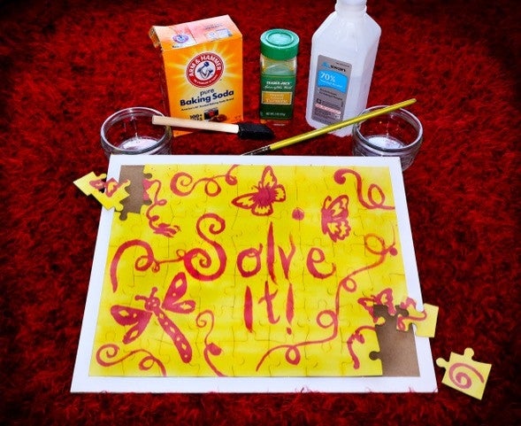 A yellow puzzle on a white board has yellow pieces with colorful red designs such as butterflies and swiggles, there’s text that says “Solve it”. The puzzle is sitting on a fuzzy red carpet, also, paint brushes, cups of water, and baking soda and other chemicals are on the table next to the puzzle. 