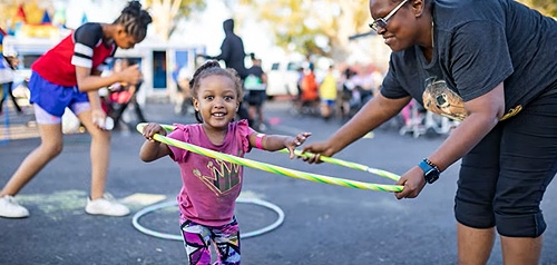 Child smiling while being taught how to use a hula hoop by an adult.
